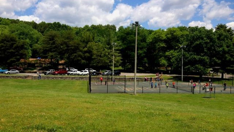 Sunny park with children playing on a playground, surrounded by trees and parked cars.
