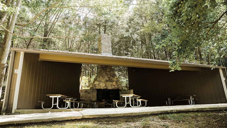 Outdoor pavilion with picnic tables and a stone fireplace, surrounded by trees.