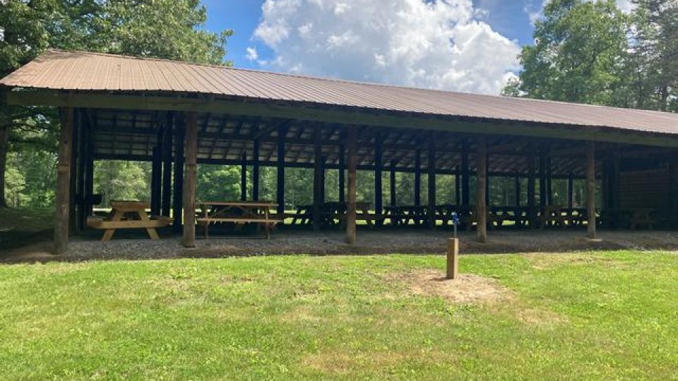 Shelter with picnic tables on grassy field under blue sky with clouds.