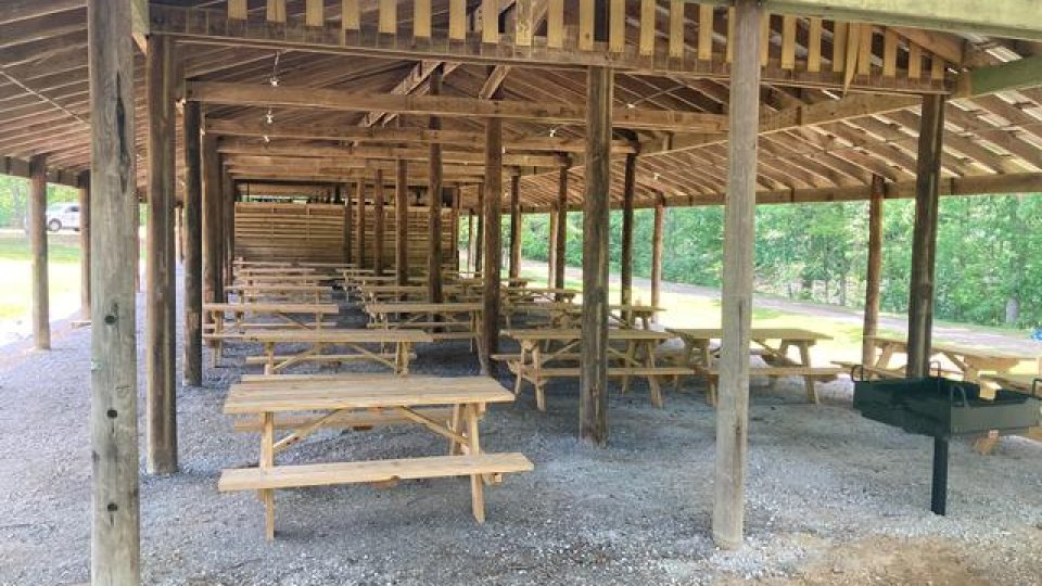 Wooden picnic shelter with empty tables and a grill, surrounded by trees.