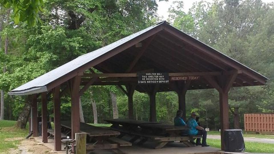 Open wooden pavilion in a park with picnic tables and trees.