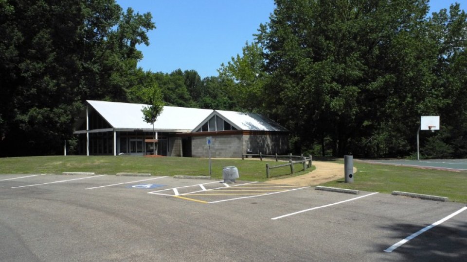 Parking lot with empty spaces, building, trees, and basketball hoop under a clear blue sky.