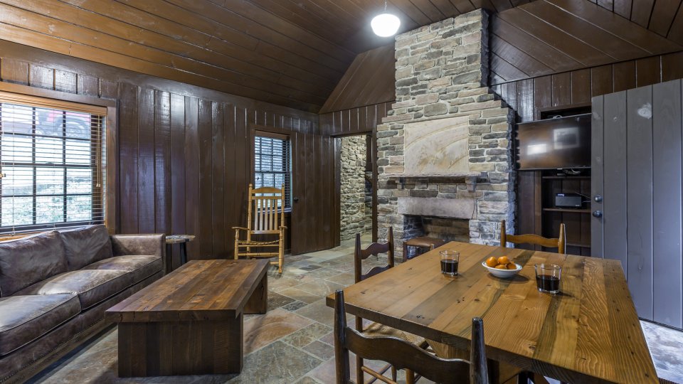 A collage featuring a small stone cabin surrounded by autumn trees, a road curving past historic stone buildings and arches, and a rustic cabin interior with wood-paneled walls, a stone fireplace, and simple wooden furniture.