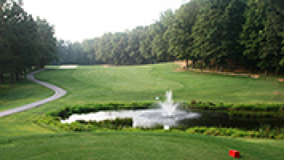 Golf course with pond and fountain, surrounded by trees.