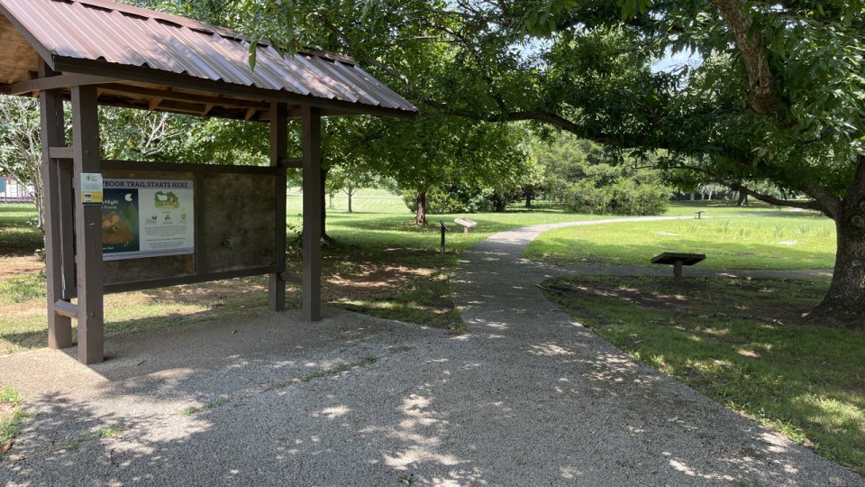 A paved trailhead is shaded by a large tree on the right. It's branches stretch across to meet the top of the trailhead kiosk. Much of the background is green with several trees and lush green, mowed grass. 