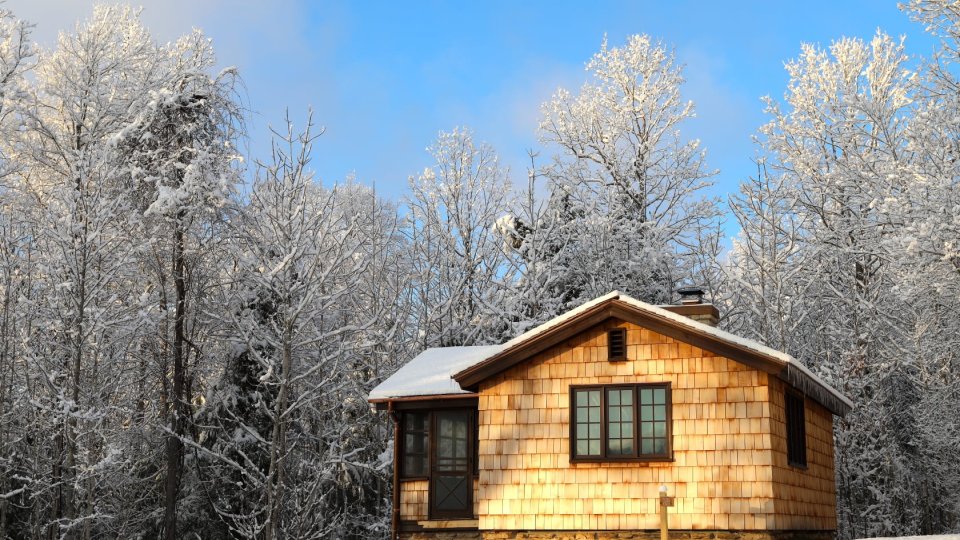 A collage showing a rustic cabin interior with wood walls, exposed beams, large windows, and a stone fireplace, alongside an exterior view of a small wood‑and‑stone cabin under a bright sky with scattered clouds.