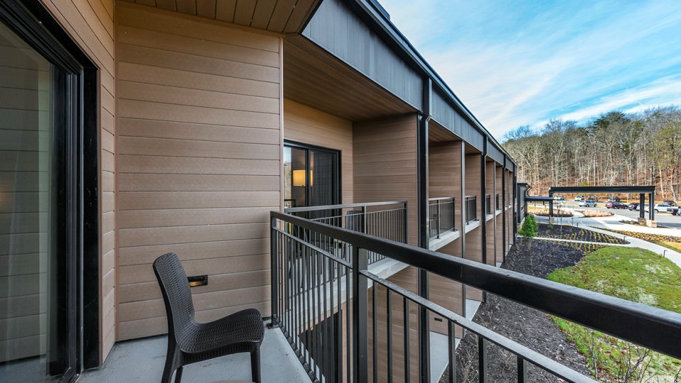 Outdoor balcony with a chair and table, overlooking a parking lot and wooded area.