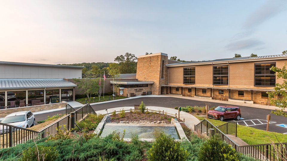 Lodge building with a parking area and fountain, surrounded by greenery at dusk.