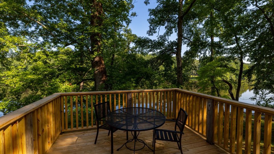Wooden deck with a small round table and two chairs, surrounded by lush trees and overlooking a calm river in sunlight.
