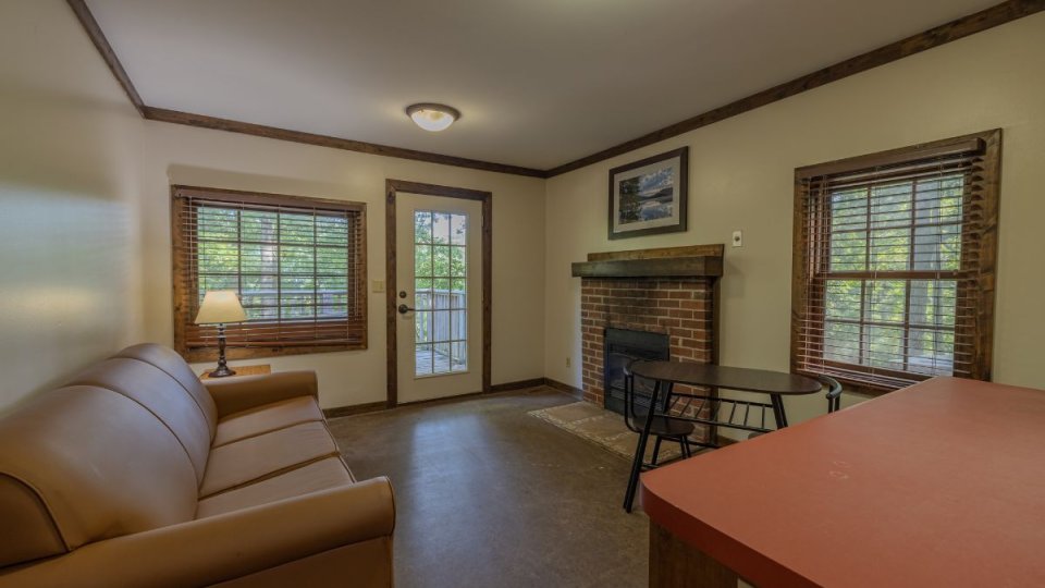 Cozy cabin interior with sofa, brick fireplace, small table, and windows overlooking trees, plus a glass door to a deck.