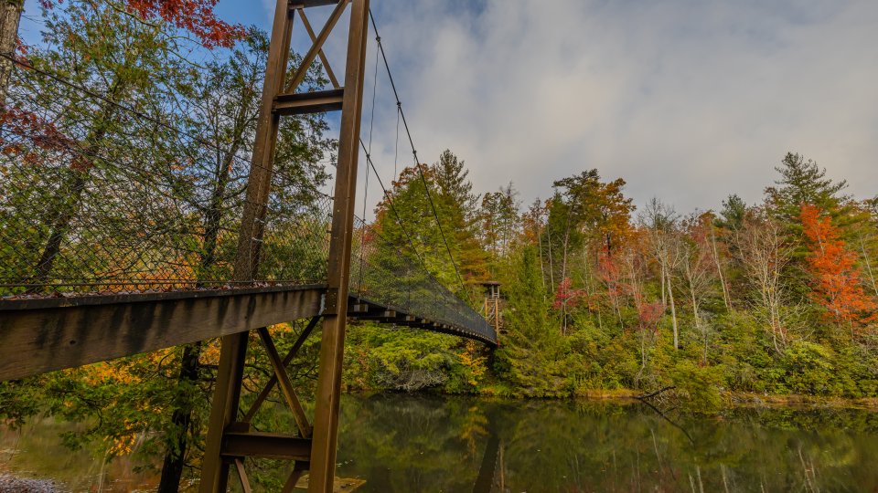Suspension bridge over a calm lake surrounded by colorful autumn trees under a partly cloudy sky.