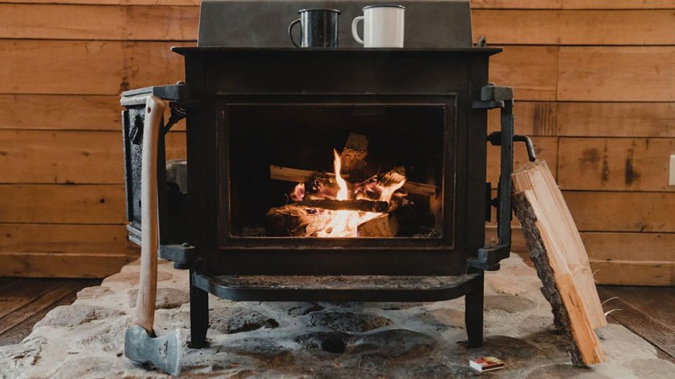 Wood-burning stove inside a cozy cabin, with a fire glowing behind the glass door. An axe leans against the stove, split logs sit nearby, and two mugs rest on top against a wooden wall backdrop.