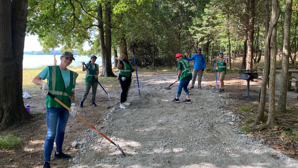 Six volunteers in green vests rake gravel on a lakeside trail under trees, smiling during a park cleanup.