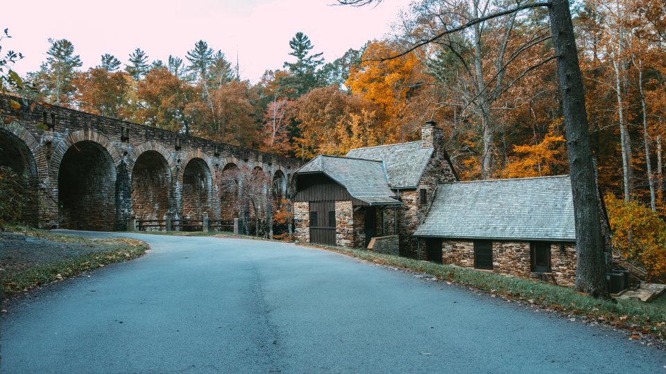 A collage featuring a small stone cabin surrounded by autumn trees, a road curving past historic stone buildings and arches, and a rustic cabin interior with wood-paneled walls, a stone fireplace, and simple wooden furniture.