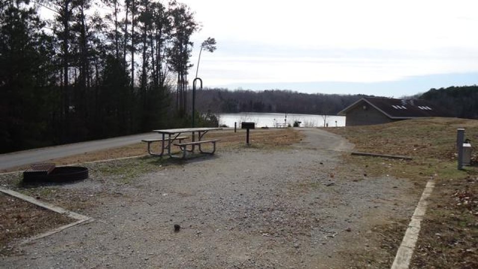 A serene campsite in winter features a grill, fire pit, and picnic table. Several trees are just to the left of the site while the lake glitters just behind it.