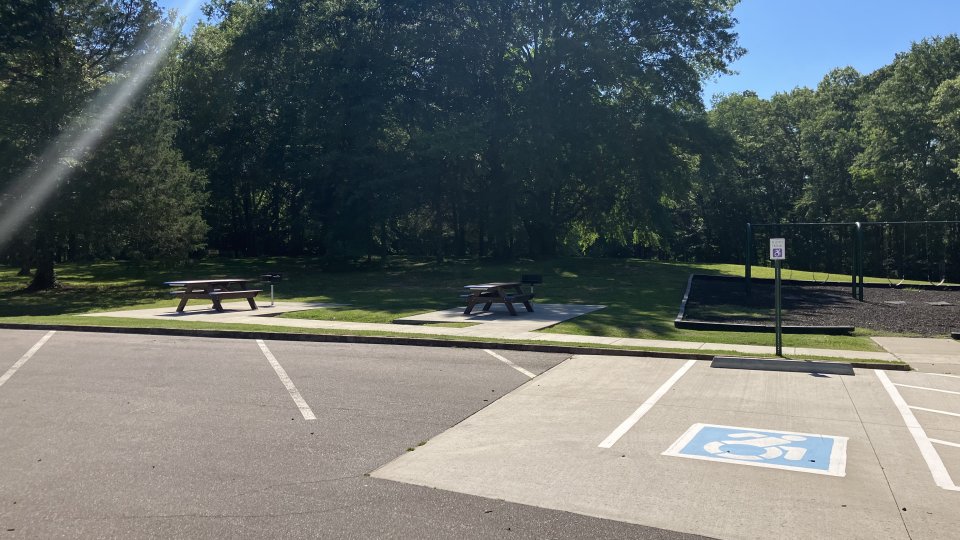 A serene park area featuring picnic tables under large trees, with a nearby playground and accessible parking spaces. Sunlight filters through the leaves.