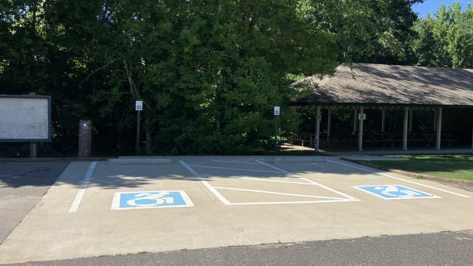 Accessible parking spaces are marked in a paved lot next to a shaded pavilion surrounded by trees. A bulletin board is visible nearby.