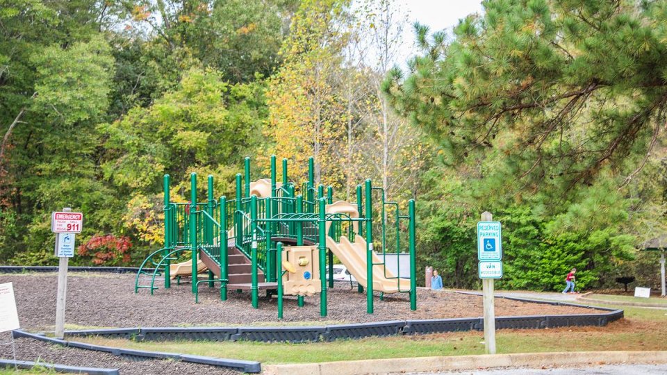 A plaground is surrounded by rubberized mulch. Trees, full of green leaves, line the background. An ADA parking space is visible in front of the play area.