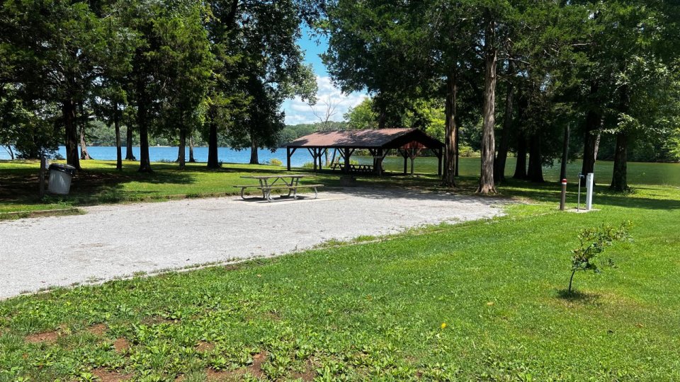 A serene park scene featuring a gravel campsite with a picnic table, surrounded by lush green grass and tall trees, overlooking a calm lake.