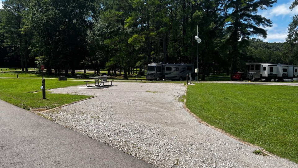 Gravel campsite surrounded by lush green grass and tall trees, featuring picnic tables and RVs parked nearby under a clear blue sky.