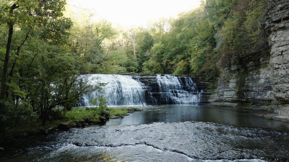 A cascading waterfall drops into a shallow pool surrounded by green trees