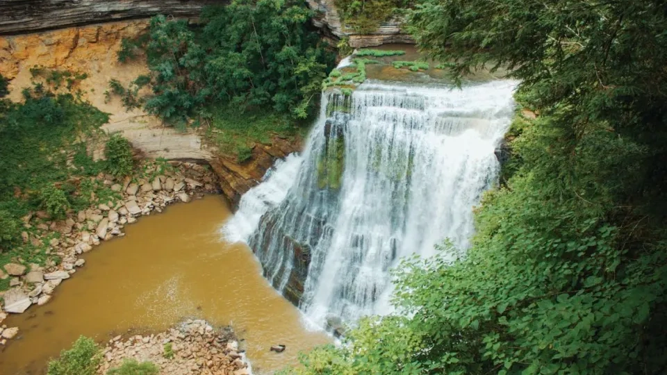 A wide waterfall cascades over layered rock cliffs into a muddy pool below, surrounded by dense green vegetation.