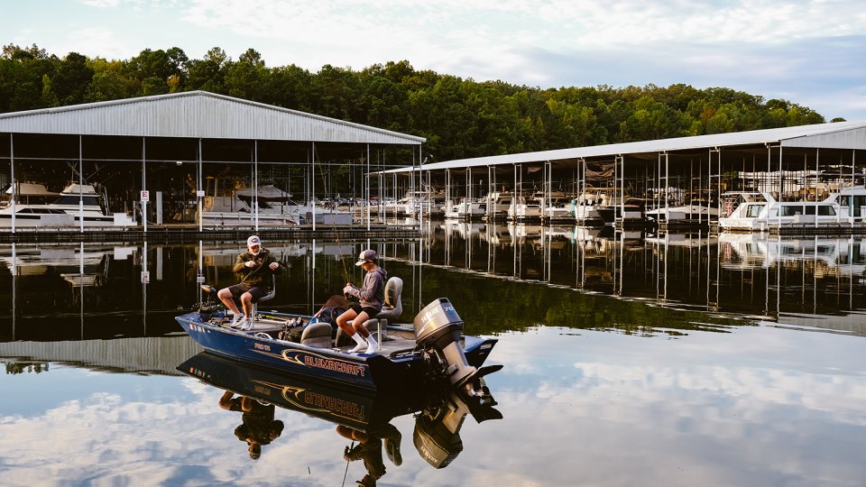 Two people fishing on a lake in a small boat near covered docks.