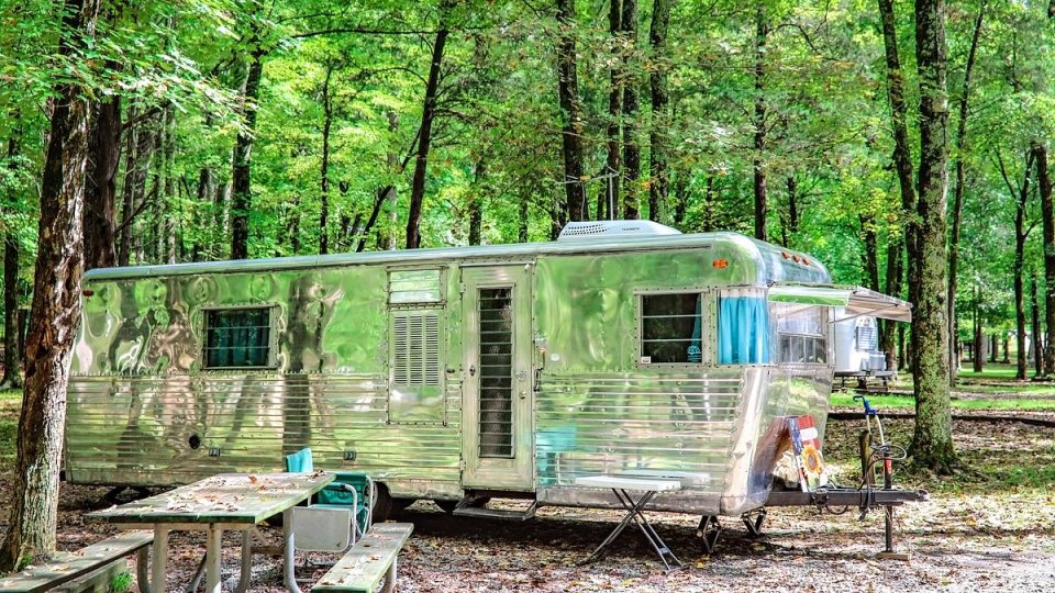 Silver trailer in a lush forest with a picnic table nearby.