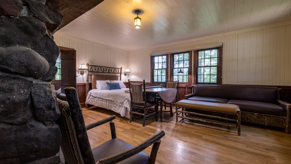Rustic cabin bedroom with log-style bed, stone fireplace, dining table, and leather sofa.