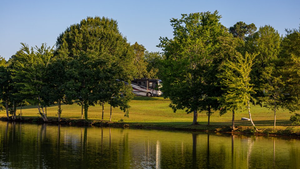 RV at a campsite among trees by a calm lakeshore with reflections in the water.