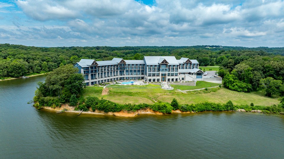 Large building by a lake, surrounded by lush green trees, under a partly cloudy sky.