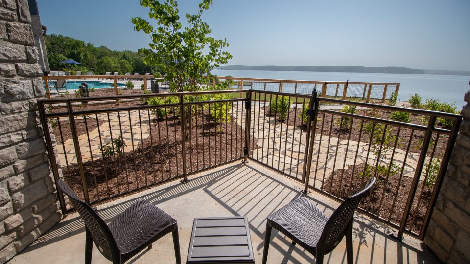 Two chairs and a table on a balcony overlooking a lake and garden under a clear blue sky.