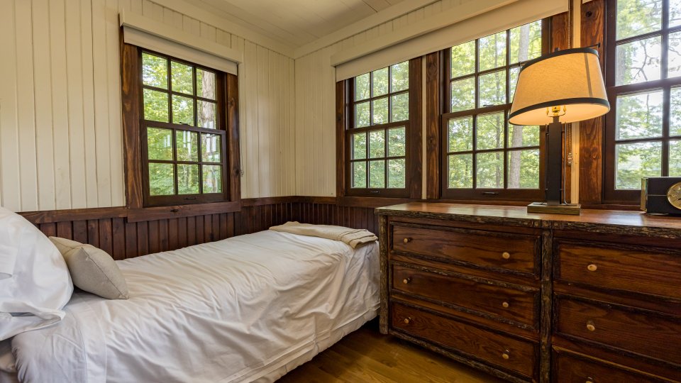 Small bedroom with a single bed, large windows, and a wooden dresser with a lamp.