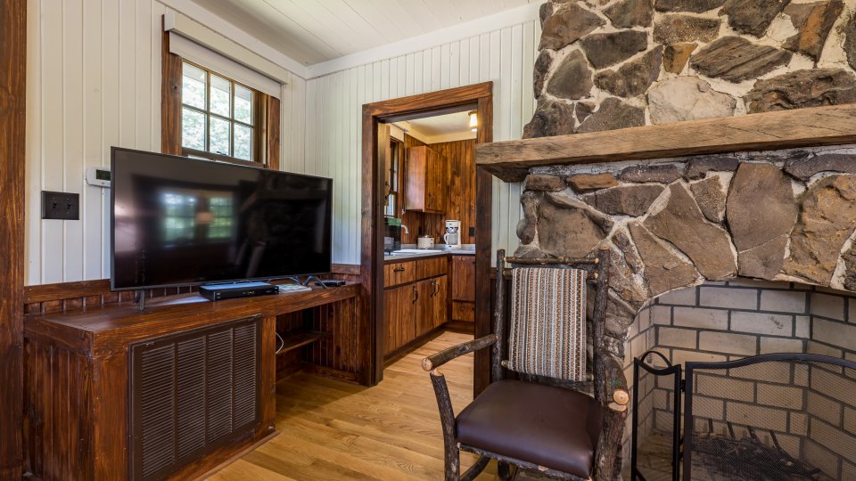 Corner of the cabin showing a stone fireplace, wooden chair, TV on a stand, and doorway into the kitchen.