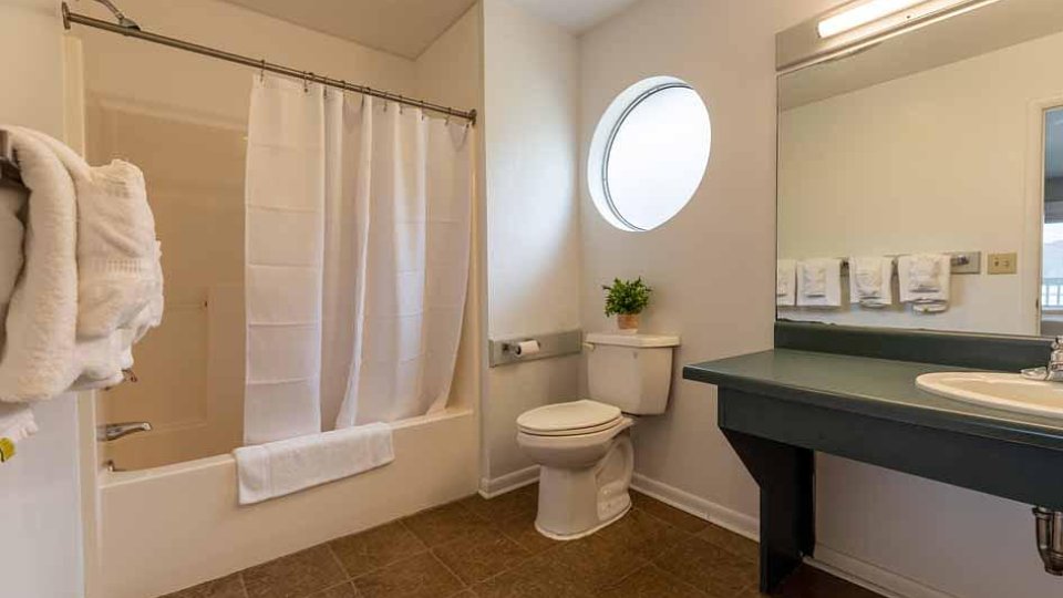 A bathroom with a tub, toilet, and sink is softly lit by a small, circular window on the wall. 