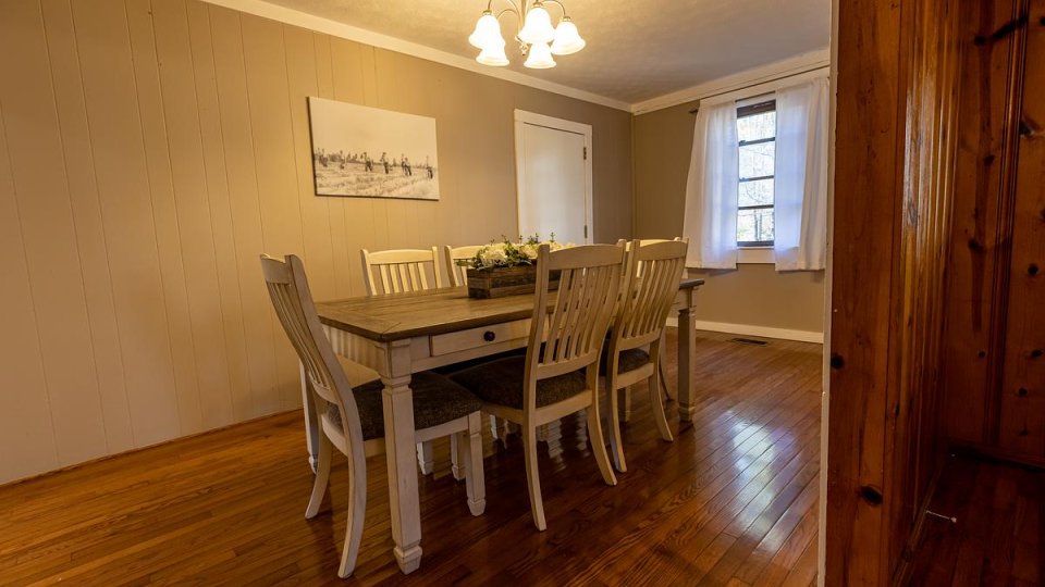 Dining room with a wooden table and eight chairs, neutral walls, wood floors, a ceiling light, and a window with white curtains.