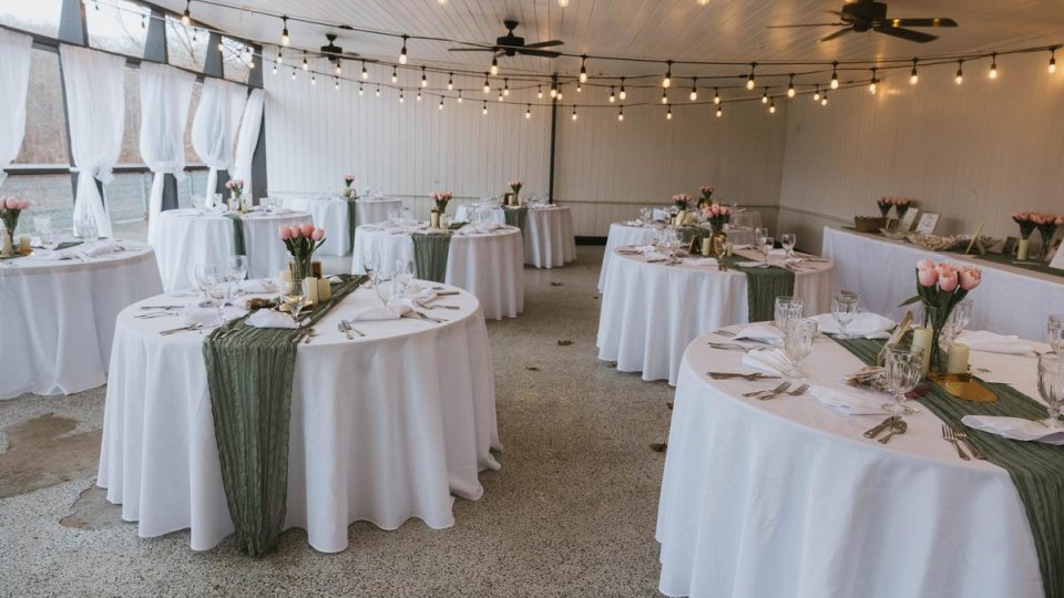 Event setup in the Lake Acorn Pavilion with round tables, white tablecloths, green runners, string lights and floral centerpieces