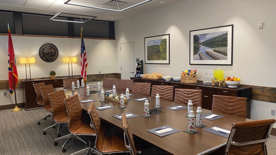Spacious conference room with a large boardroom table prepared for attendees, including reusable Tennessee State Parks water bottles, notepads, and pens. The room features geometric ceiling lights, framed nature photography, refreshments, and the Tennessee and U.S. flags.