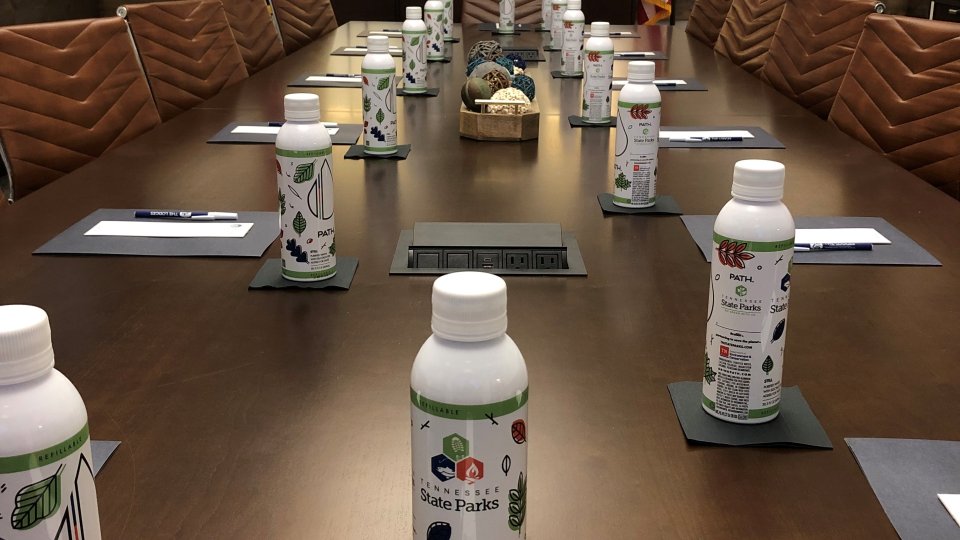 Boardroom with a long conference table set for a meeting, featuring notepads, pens, reusable Tennessee State Parks water bottles, and leather chairs. The Tennessee and U.S. flags flank the State of Tennessee seal at the front of the room.