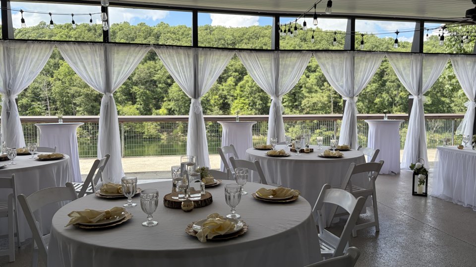 Wedding reception setup with white-draped tables, sheer curtains, string lights, and a lakeside view surrounded by trees.