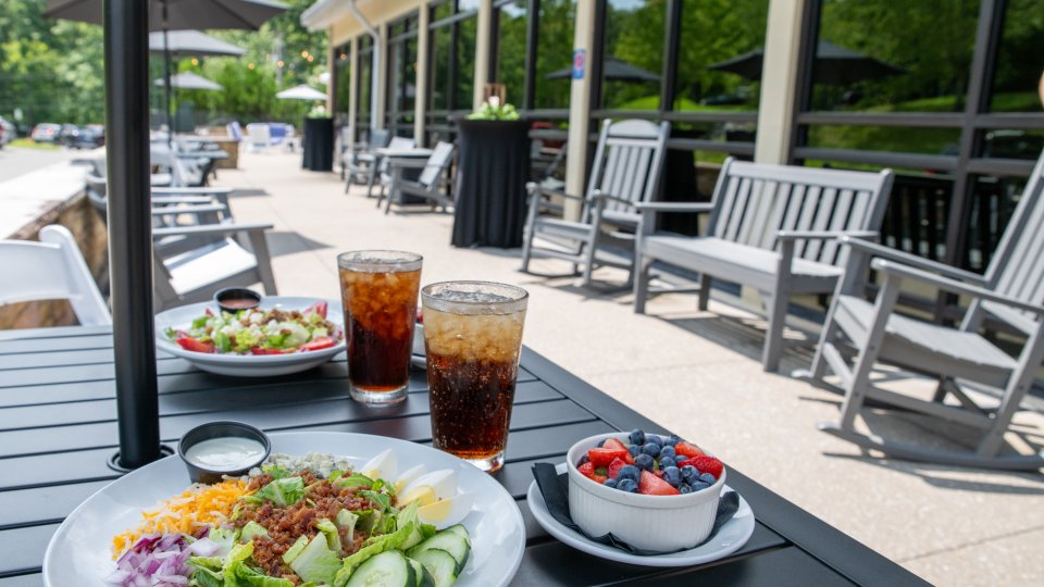 Outdoor patio table set with salads, iced drinks, and a bowl of berries beside lounge seating.