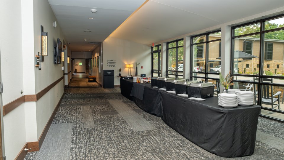 Buffet tables with food setup in a hotel corridor with large windows.