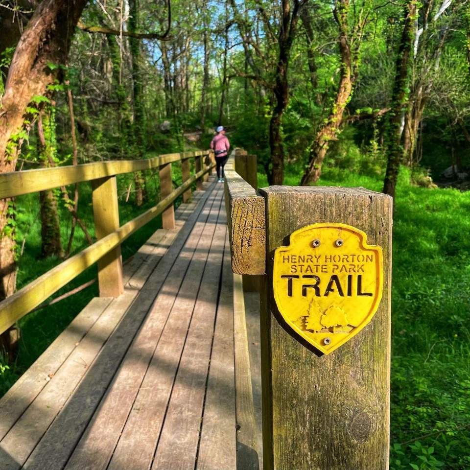 A close-up of a wooden bridge post with a yellow “Henry Horton State Park Trail” sign, looking down the bridge toward a person walking away in the distance.