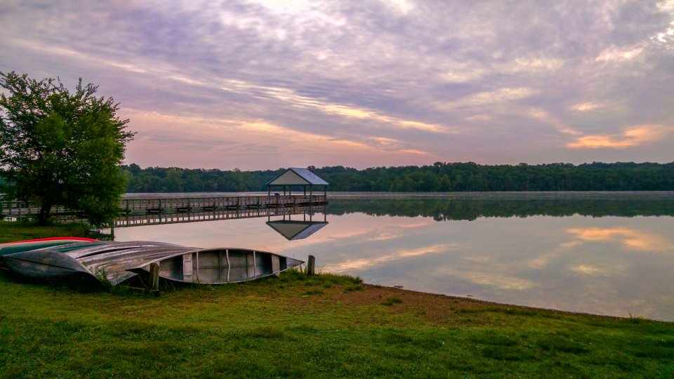 A serene lakeside scene at sunrise, two boats rest on grassy shore, with a distant dock and roofed pier.