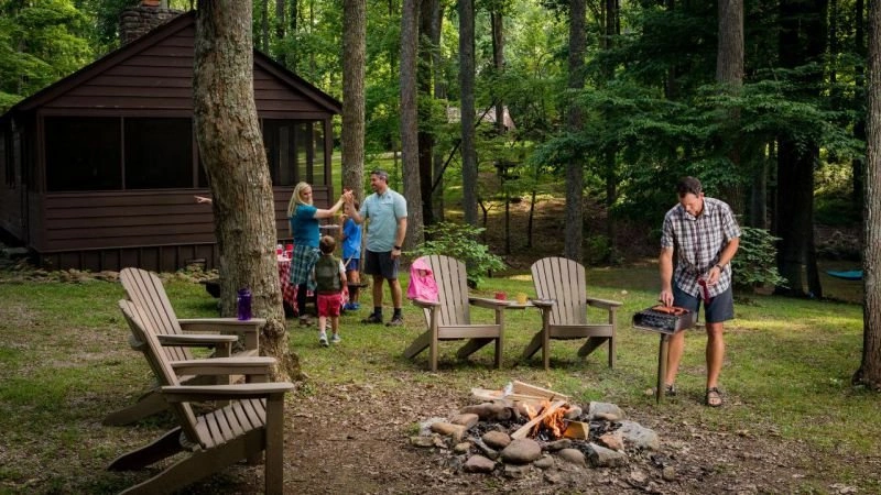 Family outside cabin in woods, man grilling by a campfire, chairs around.