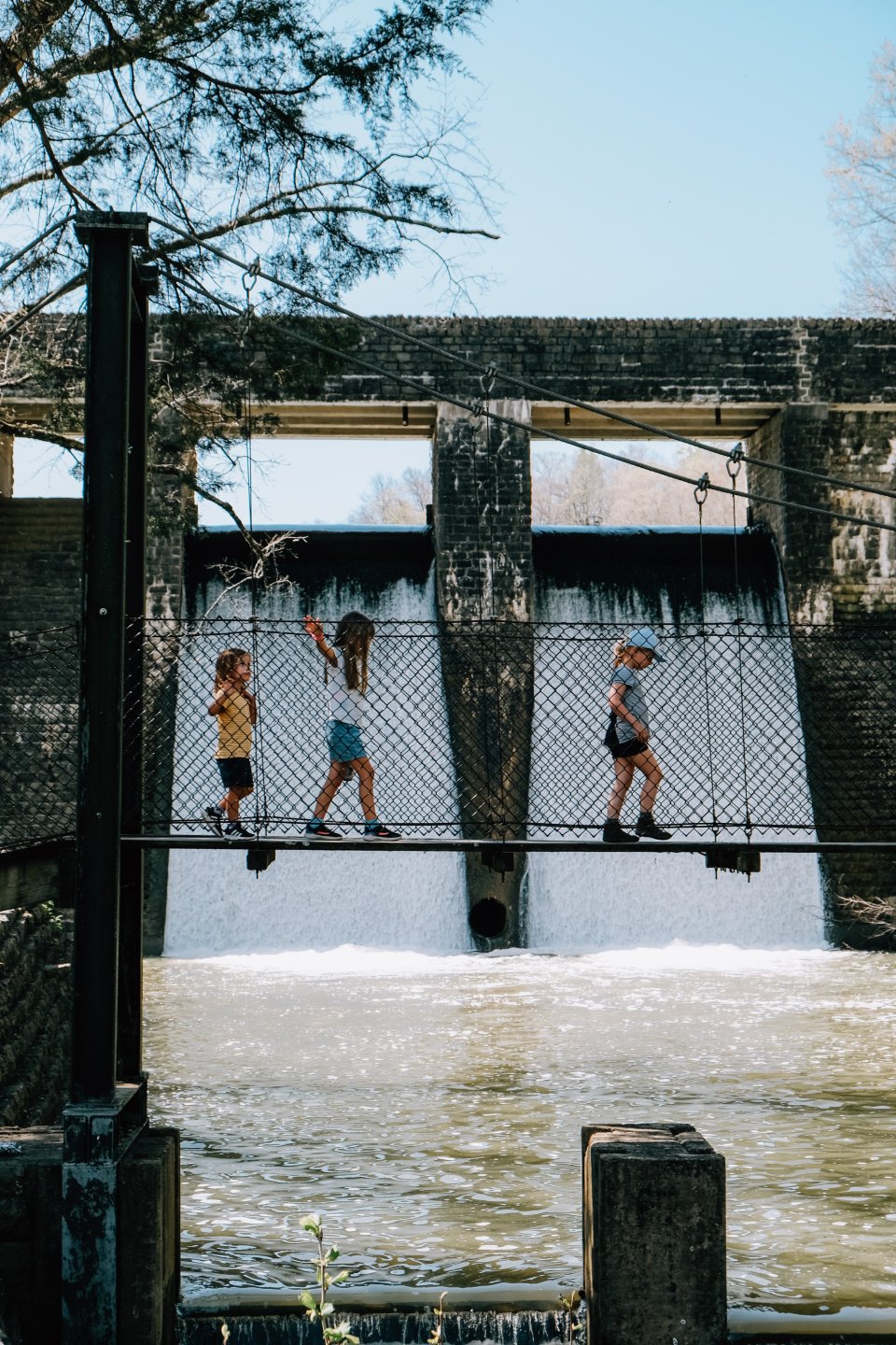 Three children walk across a narrow suspension bridge over a river, with a dam and waterfall in the background. The scene conveys adventure and playfulness.