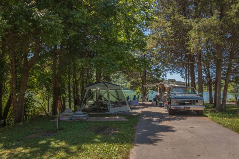 Campsite with a vintage truck and a tent under trees near a lake.