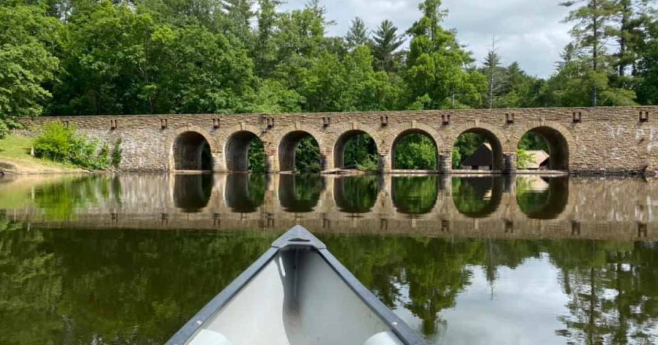 Bow of a canoe approaching a stone arch bridge over calm water.