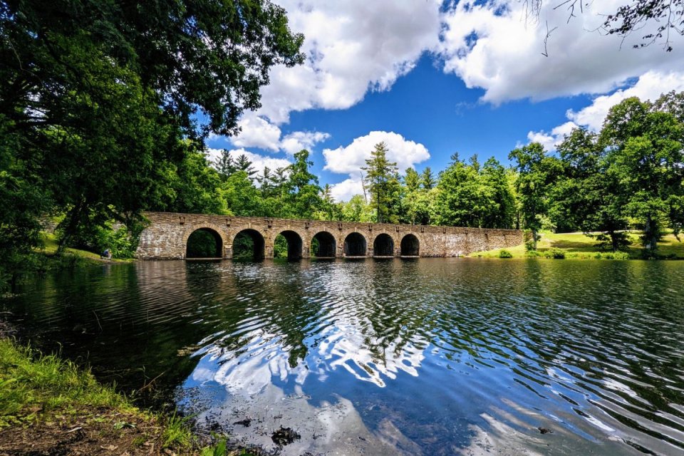 Stone bridge with multiple arches over a reflective river surrounded by lush greenery and a blue sky with clouds.