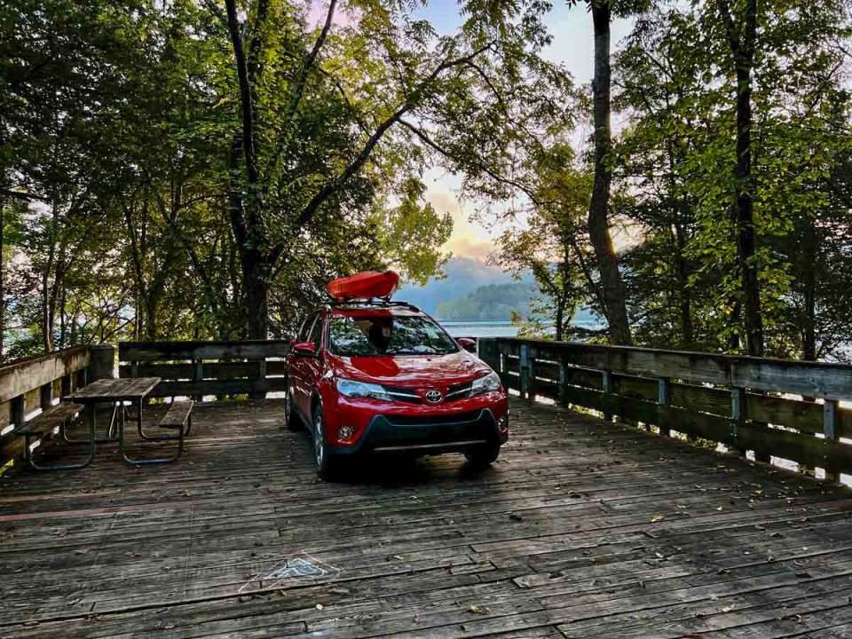 Red SUV parked on a wooded lakeside path at sunset.